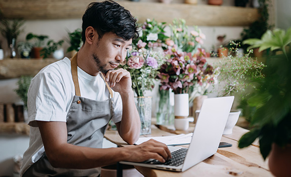 Young Asian male florist, owner of small business flower shop, working on laptop over counter 